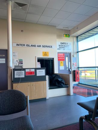 Gate 3 of Kirkwall Airport. This gate is within the main concourse, meaning no security checks at all. Next to it are seats with television showing BBC News (as usual)